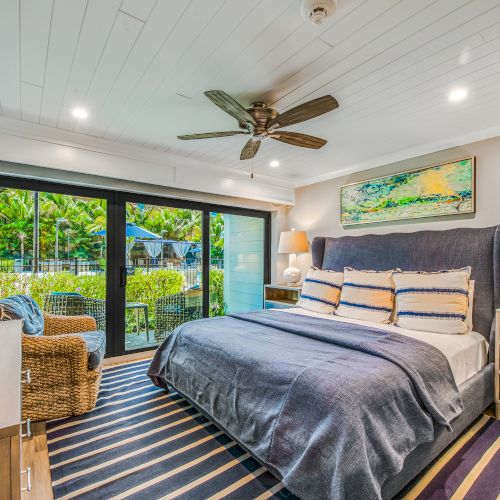 A cozy bedroom featuring a large bed with striped pillows, a wicker chair, a ceiling fan, a TV, and a glass door leading to an outdoor area.