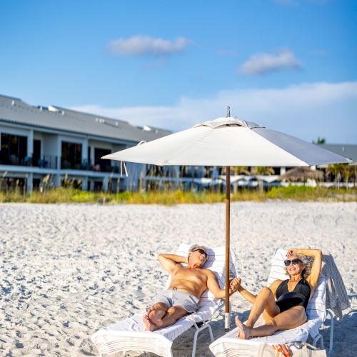 Two people lounging on beach chairs under a large umbrella on a sandy beach with buildings in the background and a clear blue sky.