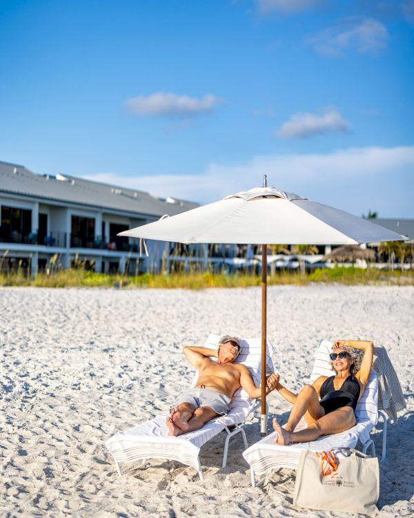 Two people lounging on beach chairs under a large umbrella on a sandy beach with buildings in the background and a clear blue sky.