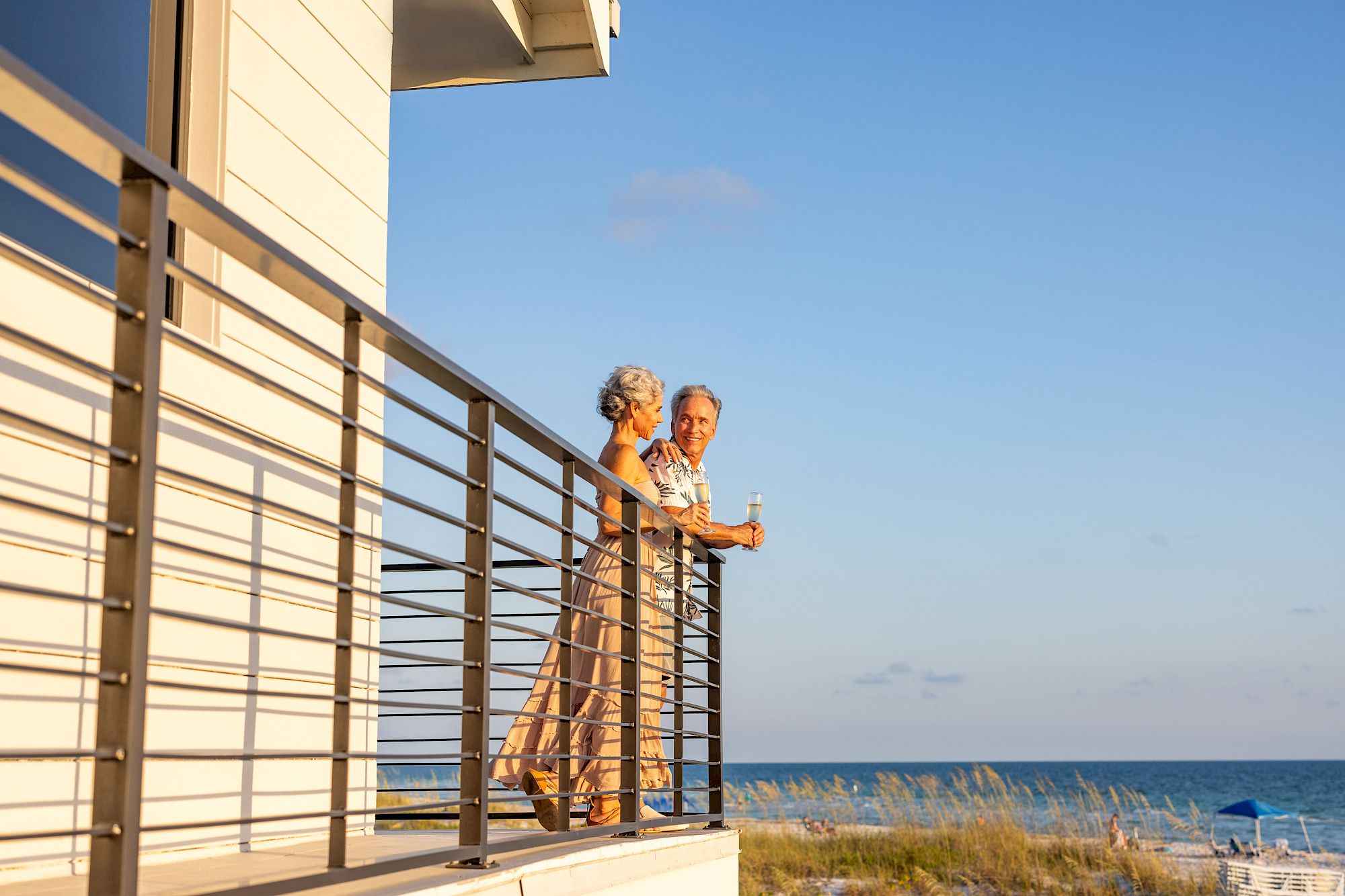 An older couple stands on a balcony by the ocean, enjoying the view and holding drinks, with blue skies and beach dunes in the background.