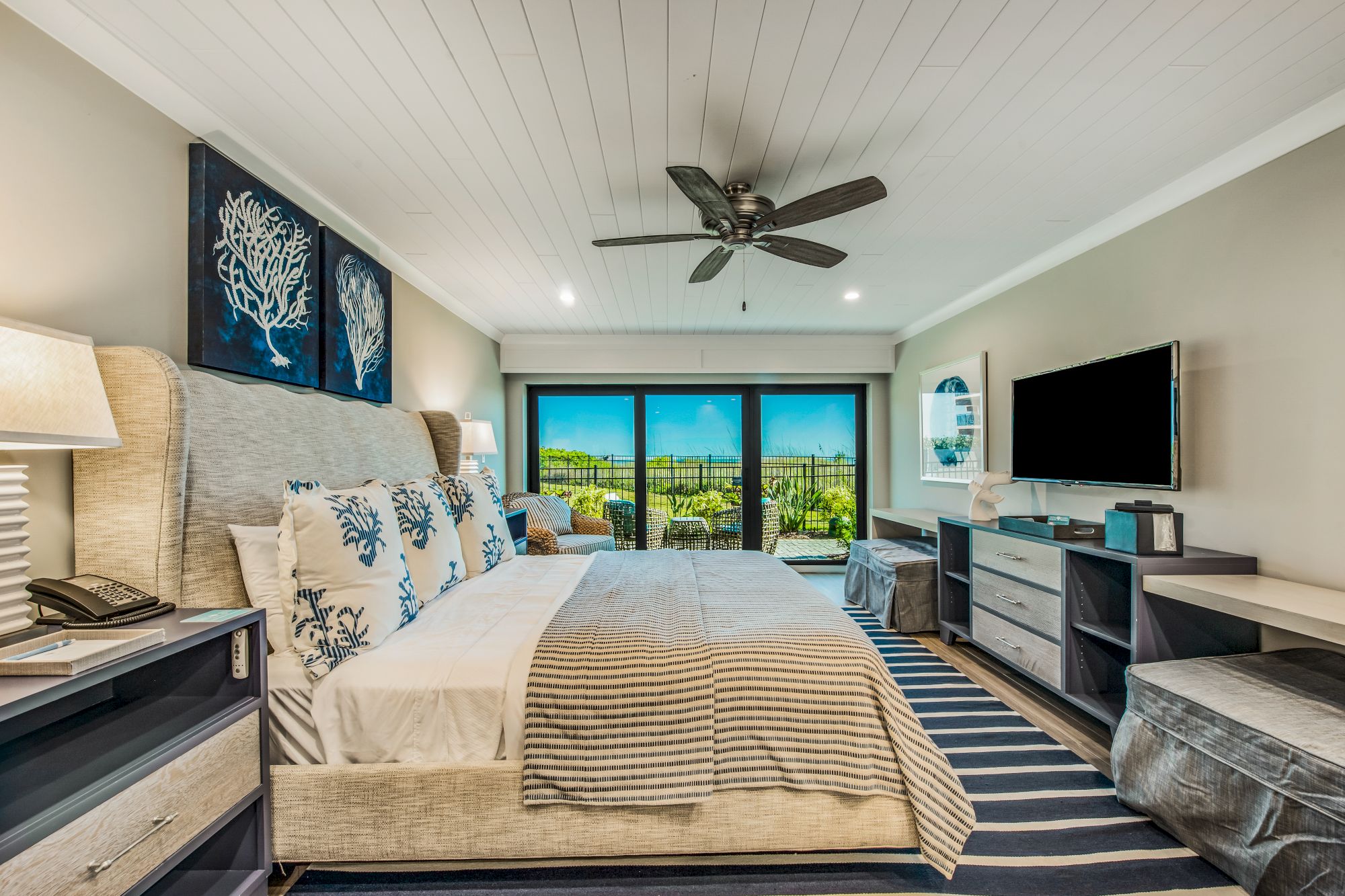 A modern bedroom with a large bed, a flat-screen TV, a workspace, and a scenic view through sliding glass doors, featuring coastal decor.