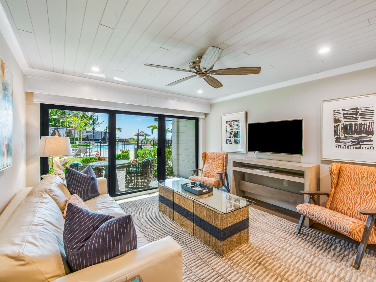 A modern living room with a white sofa, patterned chairs, a glass coffee table, a ceiling fan, a TV on the wall, and a view of a terrace through glass doors.