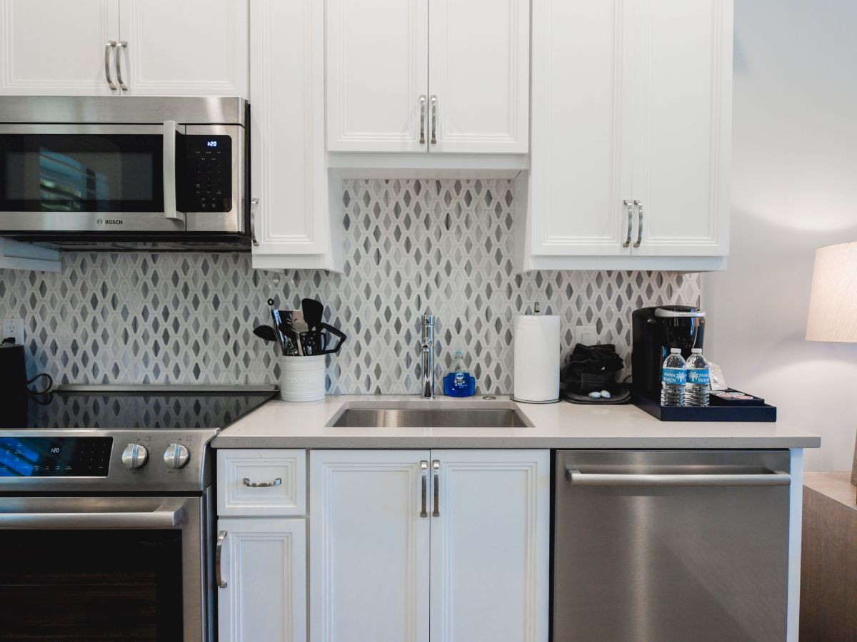 A modern kitchen with white cabinets, a sink, a microwave, a stove, a dishwasher, utensils, a coffee maker, and a lamp on the side.