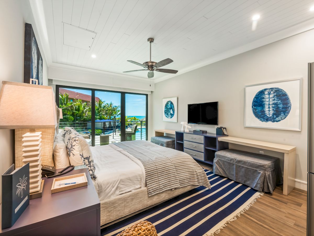 A modern bedroom with a large bed, striped rug, desk, and bench, featuring ocean-themed decor and a balcony view of greenery and water.