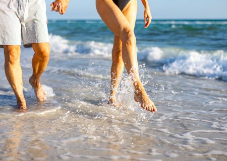 Two people walking hand in hand along the shore, with waves gently washing over their feet. One wears shorts, the other a swimsuit.