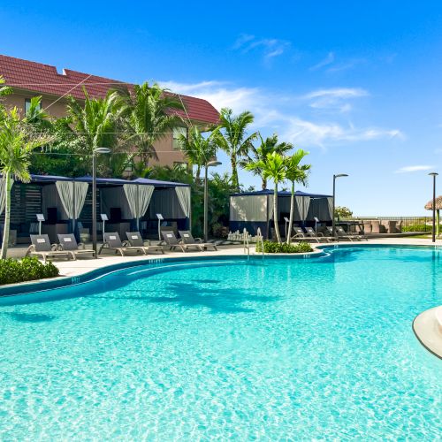 A beautiful outdoor pool area with cabanas, palm trees, lounge chairs, and a clear blue sky, perfect for relaxation and enjoying sunny weather.