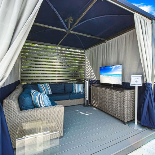 An outdoor cabana with sofas, pillows, a TV, and curtains, offering a comfortable relaxation space in a stylish setting.