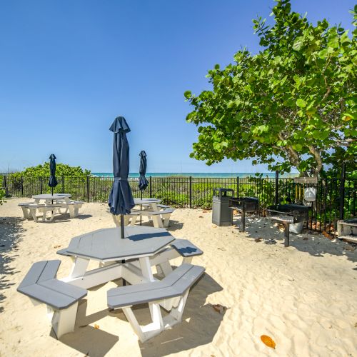 The image shows a sandy outdoor area with picnic tables, umbrellas, and BBQ grills, surrounded by greenery and a view of the ocean in the background.