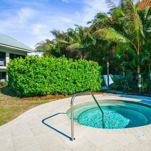 A serene outdoor hot tub with stainless steel railings, surrounded by lush greenery and hotel buildings in the background under a clear sky.