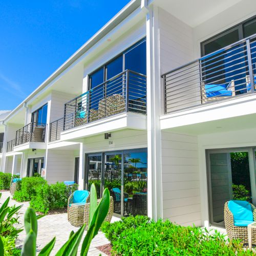 The image shows a modern, two-story building with balconies and large glass doors. There are outdoor seating areas and lush greenery surrounding the property.