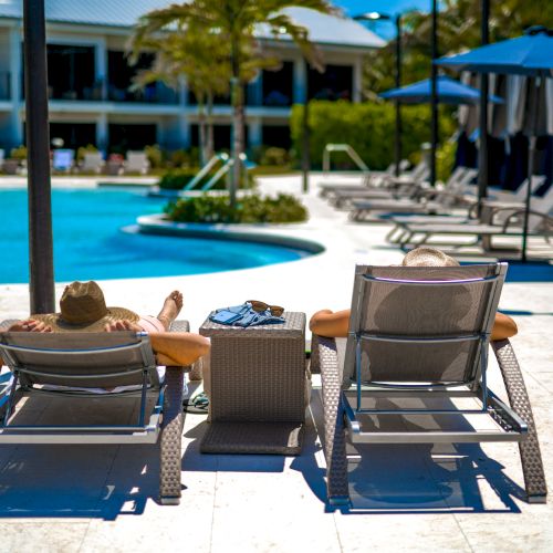 Two people relaxing on lounge chairs near a pool, with palm trees and a resort building in the background.