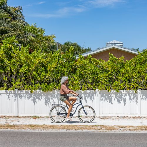 A person with gray hair is riding a bicycle on a sunny day, passing by a white fence and green foliage, with a house partially visible in the background.