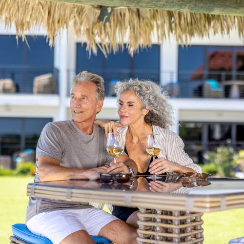 A couple enjoys a drink under a tiki-style hut, smiling and relaxed. A modern building is in the background.