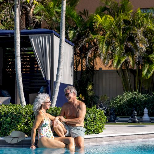 A couple is relaxing by a poolside, with the woman seated in the water and the man kneeling next to her, surrounded by tropical palm trees.