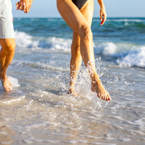 Two people, one in swim trunks and the other in a swimsuit, walk hand in hand along the shoreline as waves splash at their feet.