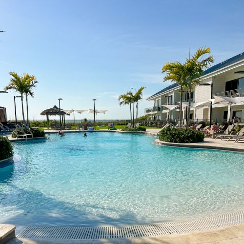 A pristine outdoor swimming pool surrounded by lounge chairs, umbrellas, and palm trees, with a view of the ocean in the background.