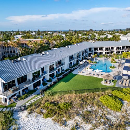 An aerial view of a resort with a large building, a pool, lounge areas, and landscaping, located adjacent to a sandy beach.