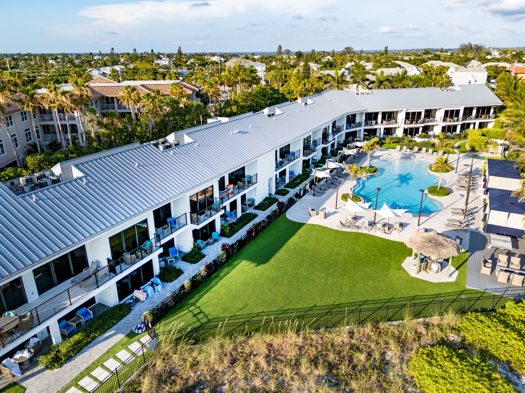 An aerial view of a resort featuring a large curved building, swimming pool, lounge chairs, umbrellas, and lush landscaping.