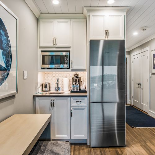 A modern kitchenette with white cabinets, stainless steel refrigerator, microwave, coffee maker, and toaster, and a framed blue artwork on the wall.