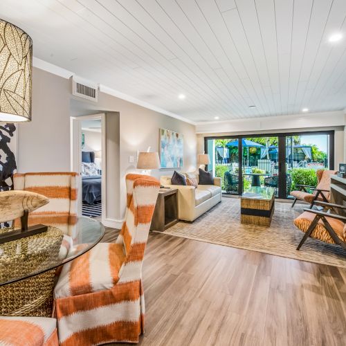 A modern living area with patterned chairs, a dining table, kitchen, and a seating area opening to a garden view through glass doors.