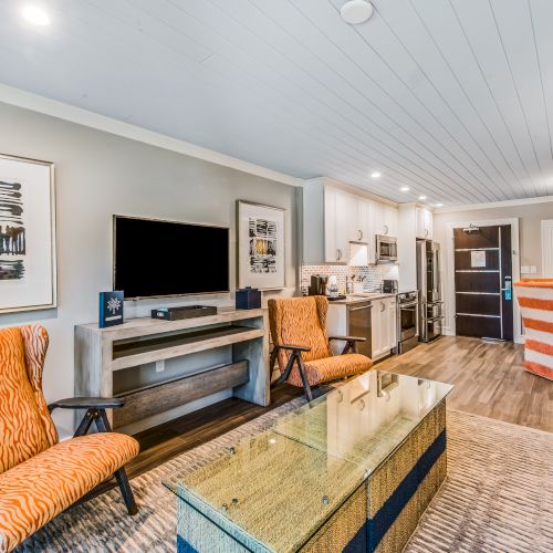 A modern living room features orange-patterned chairs, a glass table, a flat-screen TV, art on the walls, and a view of a kitchen and dining area.