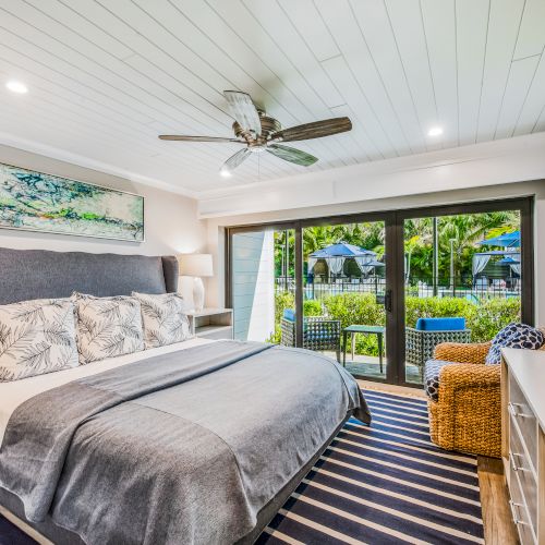 A cozy, modern bedroom with a king-sized bed, ceiling fan, large TV, and sliding doors leading to an outdoor view of greenery and a seating area.