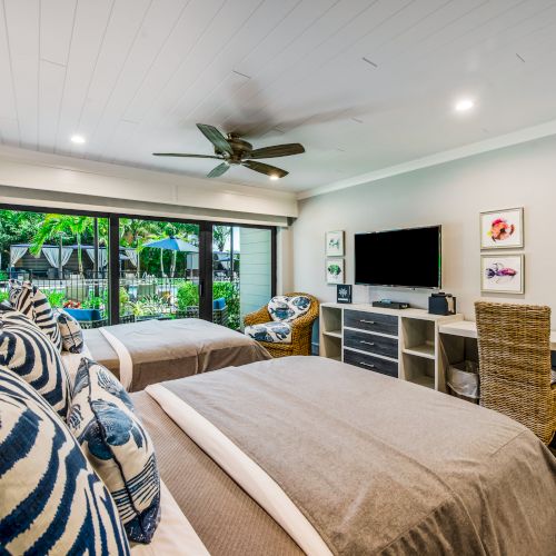 This image shows a bedroom with two twin beds, a ceiling fan, a TV on a dresser, wicker chairs, and a large window overlooking greenery outside.