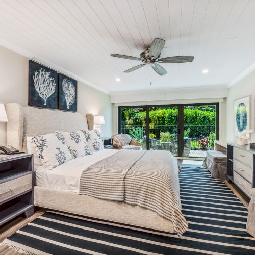 A cozy, modern bedroom features a large bed, striped rug, ceiling fan, nightstands, TV, and glass doors opening to a lush, green view outside.