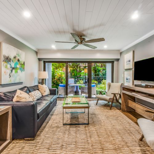 A modern living room with a sofa, TV, glass coffee table, and large sliding glass doors leading outside to a lush garden patio.