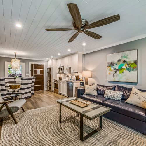 A cozy living room with a ceiling fan, modern furniture, a sofa, coffee table, abstract art, and a view of the kitchen and dining area in the background.