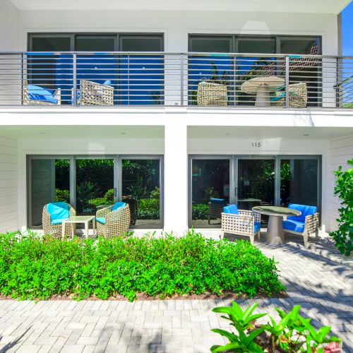 A two-story building with balconies, patio furniture, and lush greenery. The ground floor has doors leading to outdoor sitting areas ending the sentence.