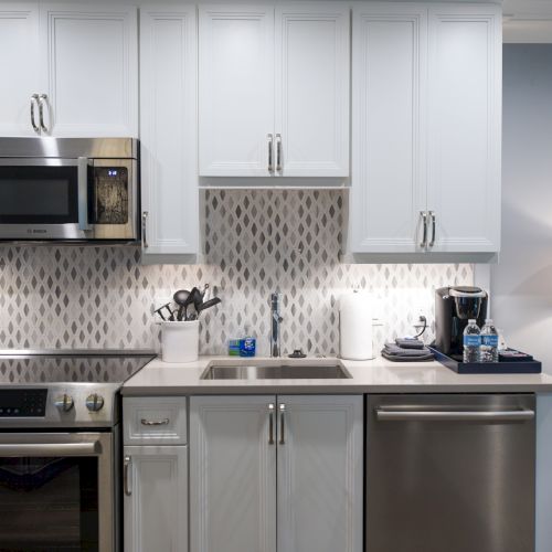 A modern kitchen with white cabinets, stove, oven, microwave, sink, dishwasher, a lamp, and other small appliances on the counter.