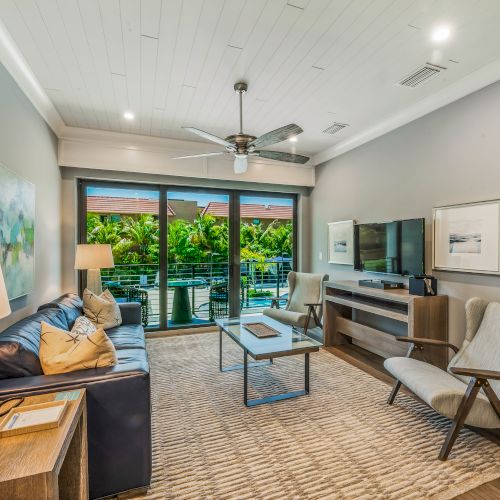 This image shows a modern living room with a sofa, armchairs, coffee table, TV on a stand, lamp, artwork, and sliding doors opening to a balcony.