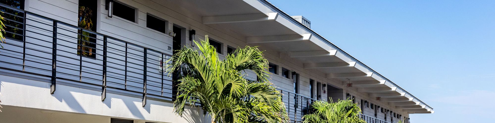 The image shows a modern, two-story building with balconies, surrounded by palm trees and greenery, under a clear blue sky.