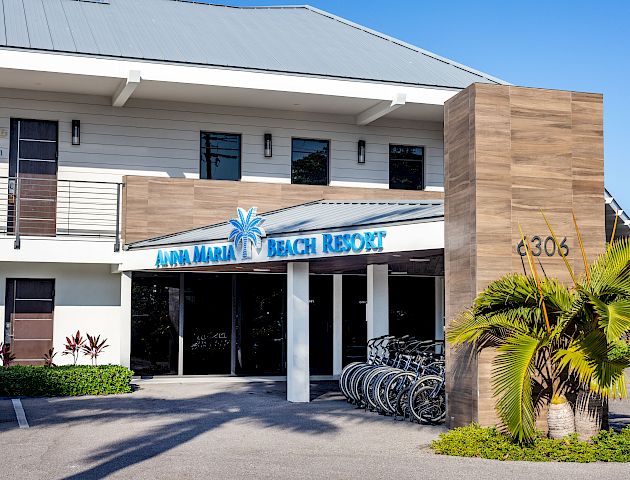 The image shows the entrance of Anna Maria Beach Resort, featuring a modern building with a sign and the address 