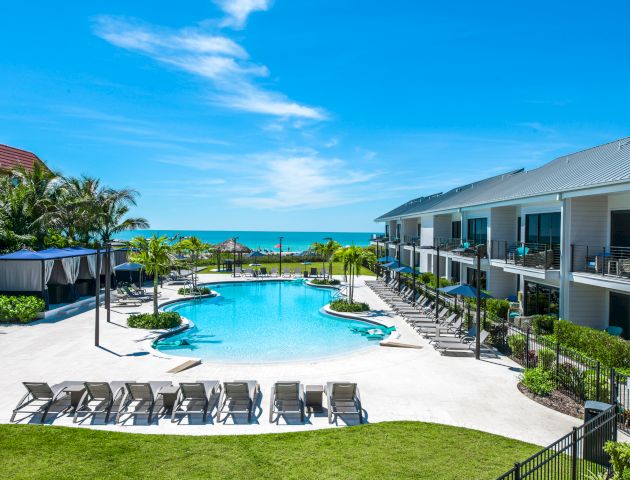 The image shows a resort with a pool, lounge chairs, palm trees, and buildings, all set against a backdrop of a clear blue sky and ocean.