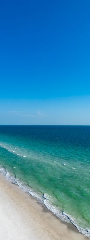 The image shows a stretch of white sandy beach meeting clear blue-green ocean waters under a clear blue sky.