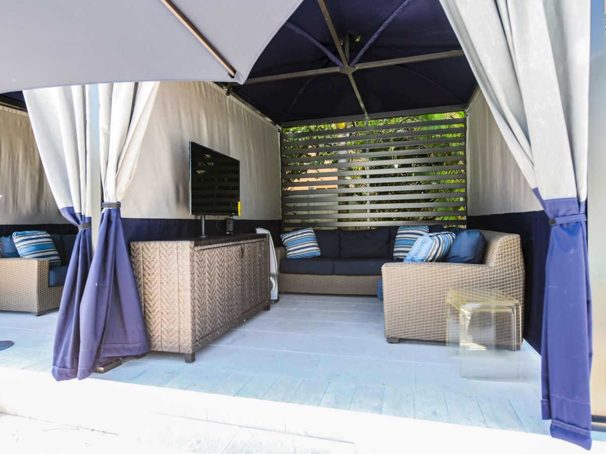 A cozy outdoor lounge area under a canopy with wicker furniture, cushions, a TV, and drapes, creating a sheltered and inviting atmosphere.