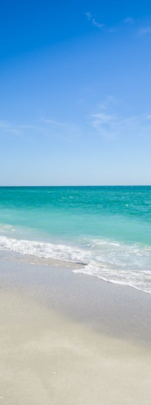 This image shows a serene beach with white sand, clear turquoise water, and a blue sky with few clouds, depicting a peaceful coastal scene.