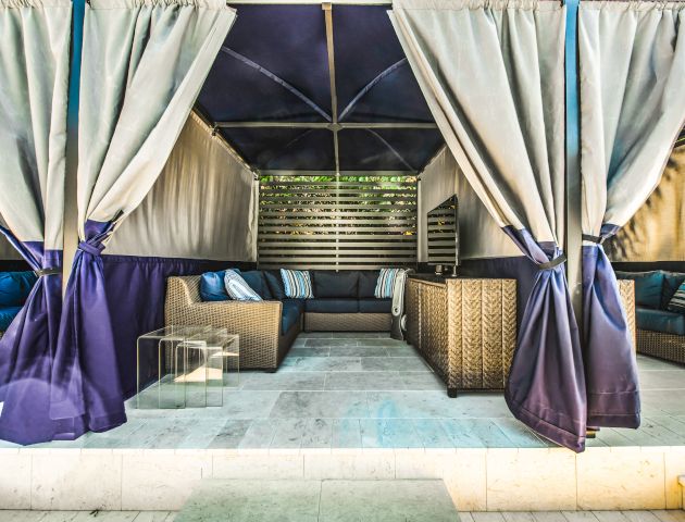 Cozy outdoor cabana with wicker furniture, blue cushions, and white drapes offering a shaded lounge area on a tiled floor.