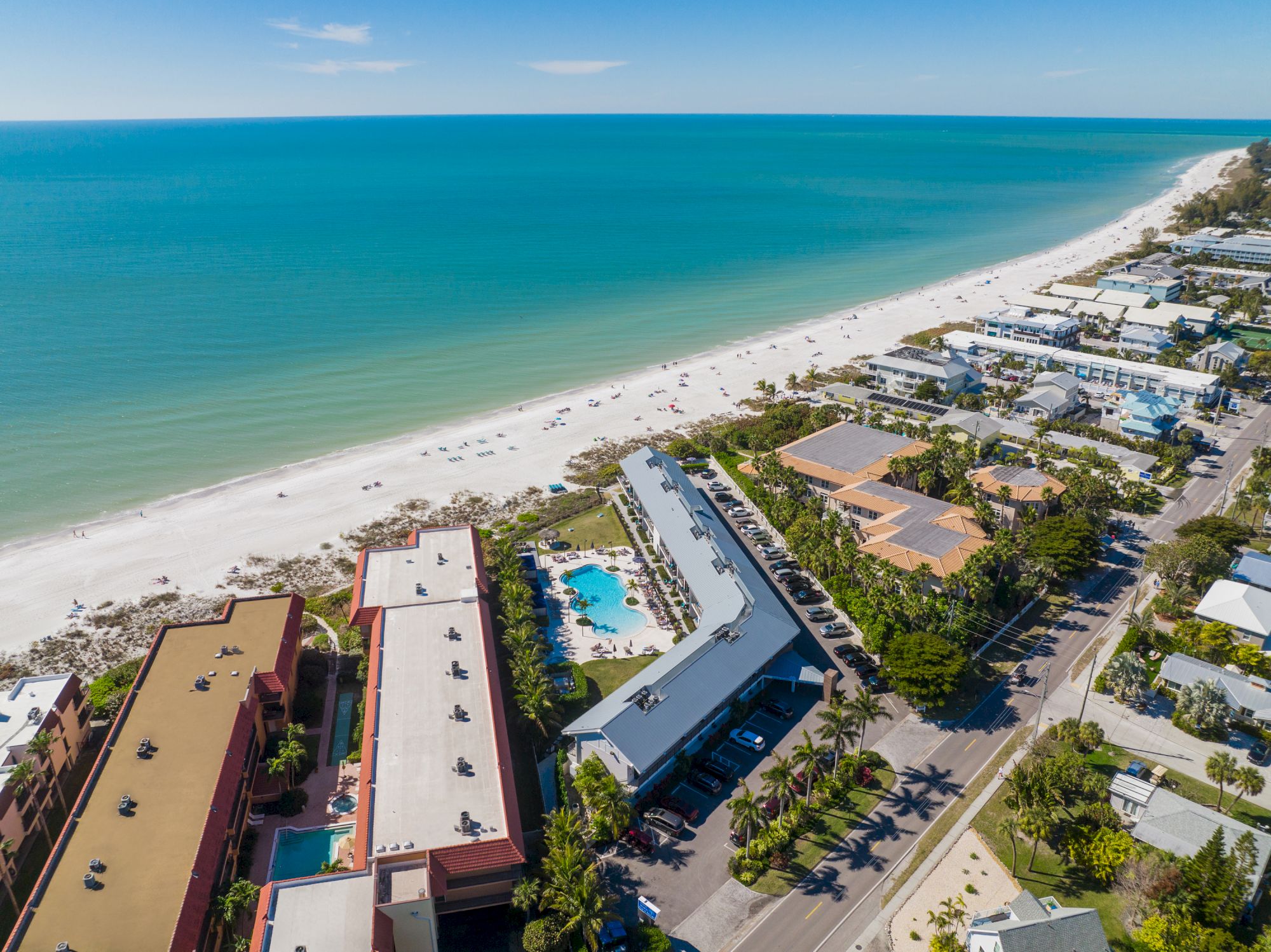 This image shows an aerial view of a beach lined with buildings and a road parallel to the shoreline, with clear blue water in the background.