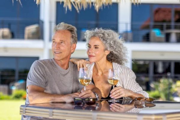 A couple is sitting at an outdoor table under a thatched roof, enjoying drinks and each other's company in a sunny setting.