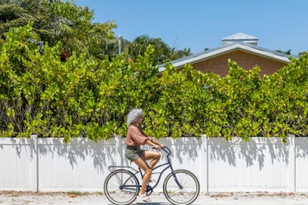 A person with grey hair is riding a bicycle along a road, passing a white fence with green shrubs and a house in the background.